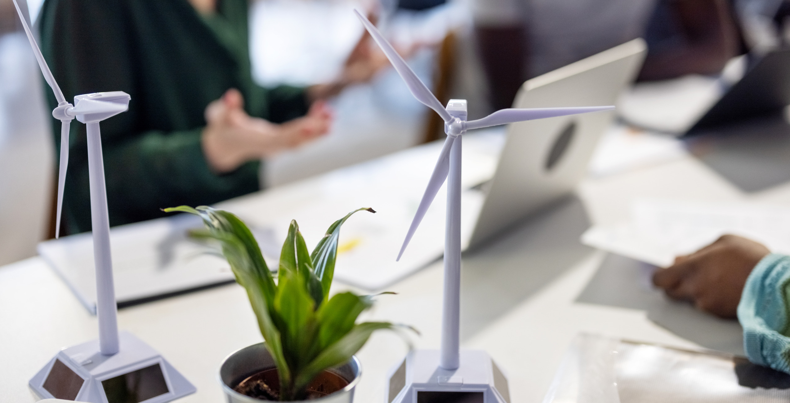 3D models of wind turbines on a desk. Small green plant in the foreground. Background is blurred suggesting two to three people with laptops.
